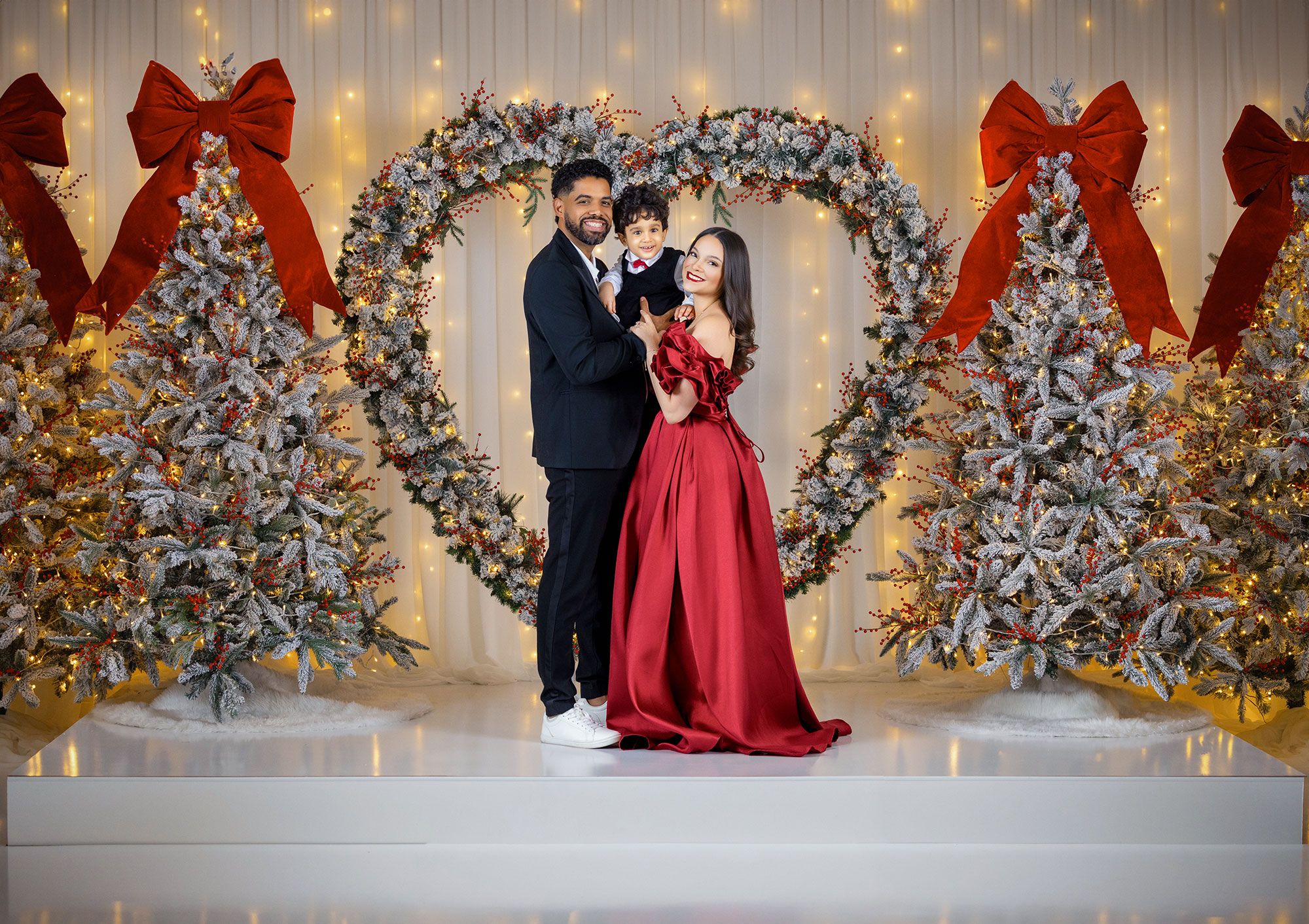 Romantic holiday photoshoot with frosted Christmas trees, red bows, and twinkling lights creating a magical backdrop.