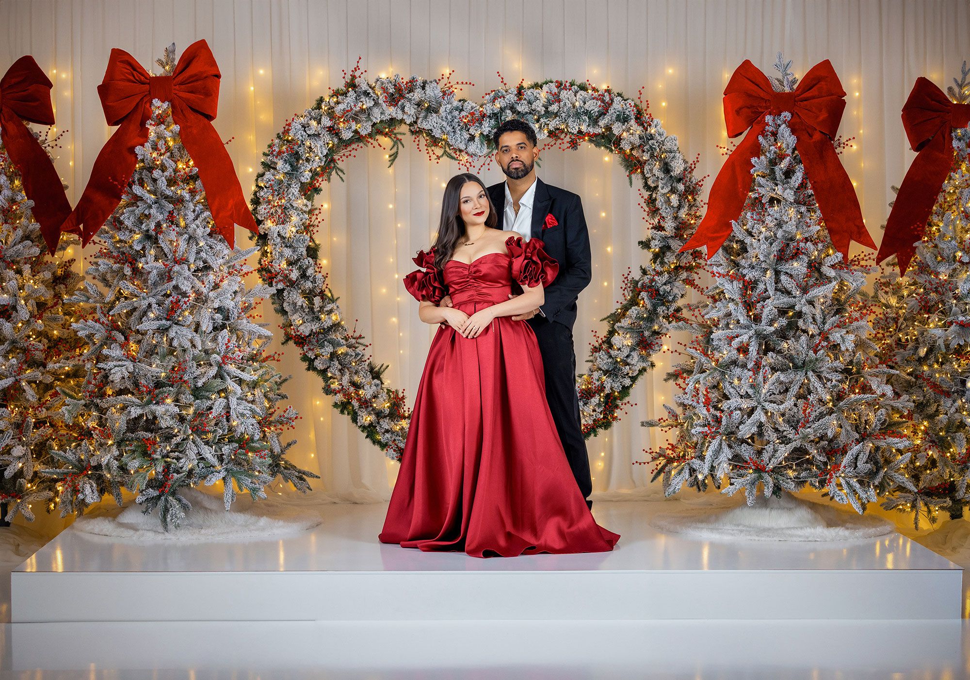 Elegant couple poses against a winter wonderland backdrop with flocked Christmas trees and red decorative bows.