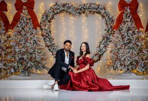 Couple in formal attire poses by decorated Christmas trees with red bows and sparkling lights.