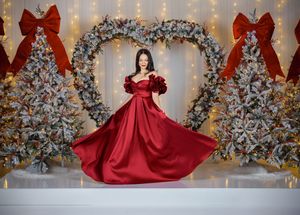 A flowing red satin gown with a bow detail is showcased against a silver and white holiday wreath backdrop.