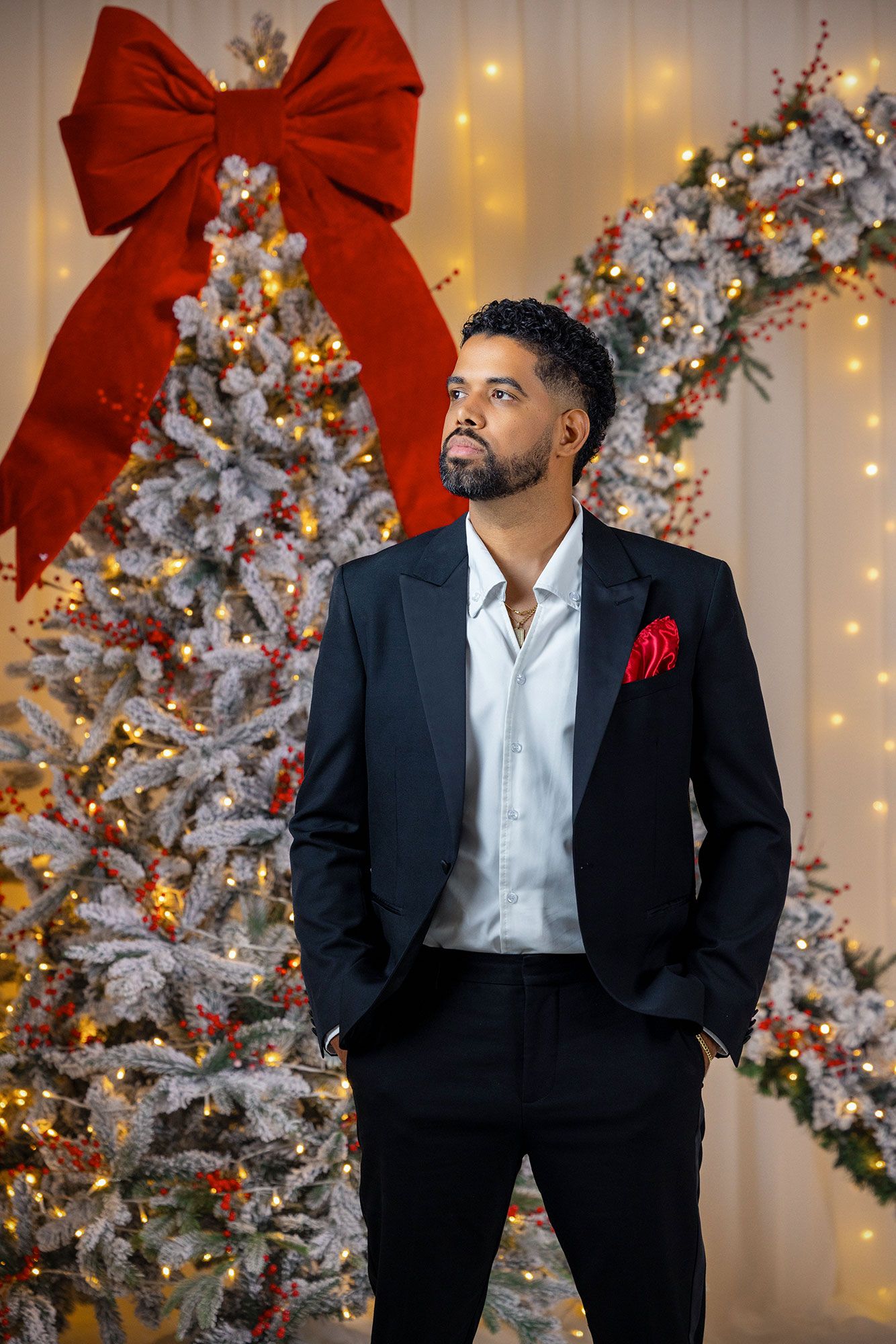 A person in a black suit and white shirt poses in front of a decorated Christmas tree with red bows and twinkling lights.