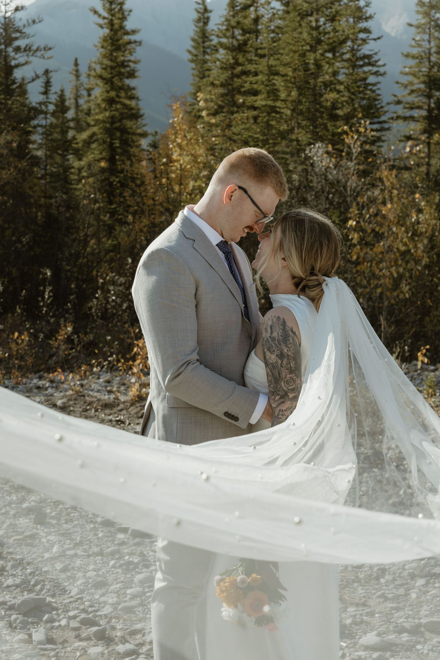 A romantic winter wedding photo series showing a couple embracing as a white veil flows dramatically in the snowy mountain setting. Minot