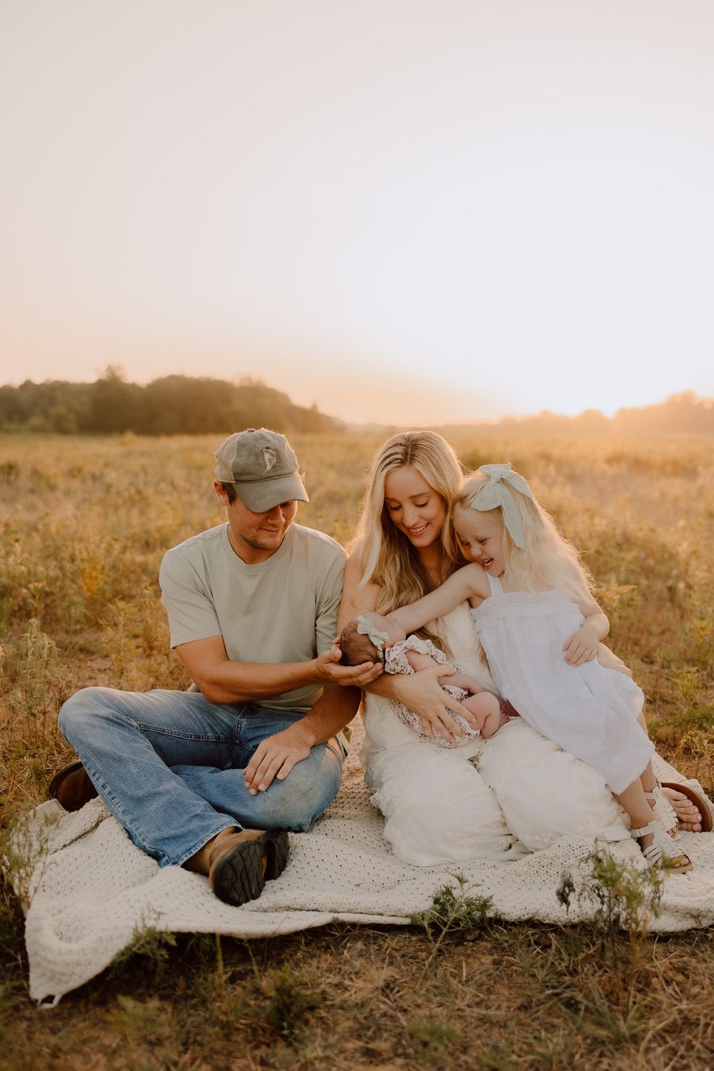 Family sits together on blanket in golden field during sunset sharing tender moment outdoors.