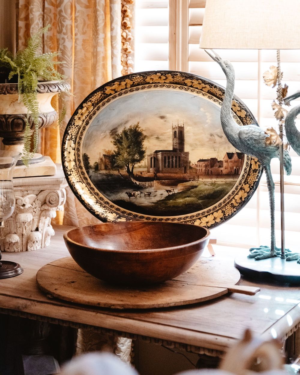 A hand-painted platter of an old village surrounded by a blue bird lamp and a vintage wooden bowl, and a cutting board.