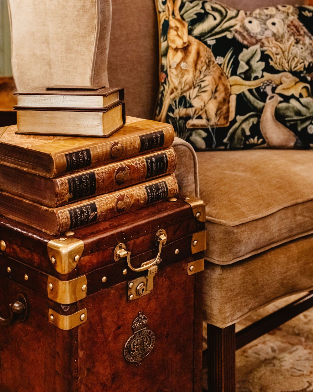 An antique chest with stacks of books sitting on top, with a tan cushioned chair, along with a wood-themed pillow