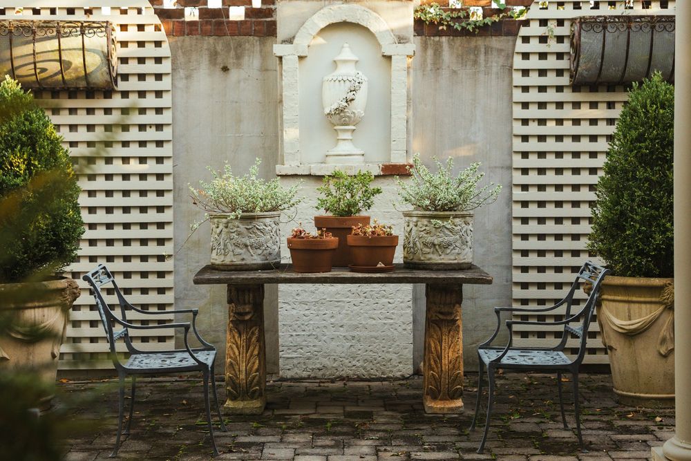 A garden table with three potted plants sits between two large planter vases with tall evergreen shrubs growing out of them