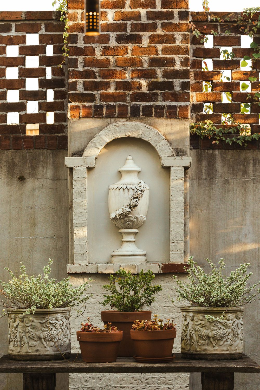 A cream sculpture of a vase carved into the garden wall. In front is a garden table with three medium-sized pots and two small pots