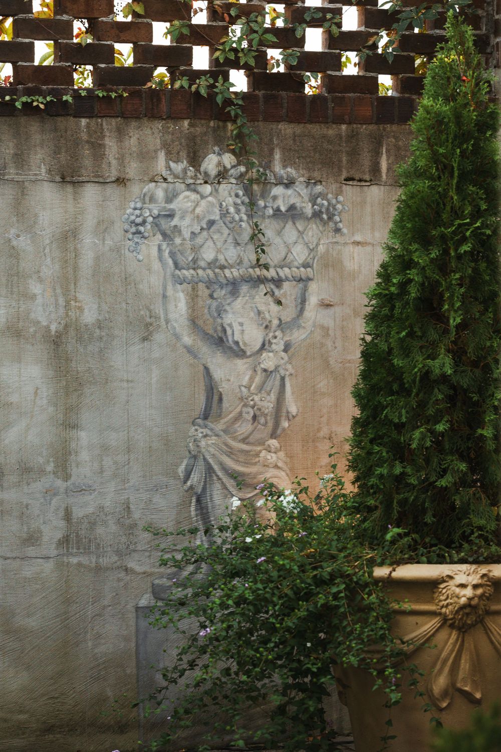 Painted mural of a woman holding a fruit basket over her head, tucked behind a large potted plant with an evergreen bush growing out of it.