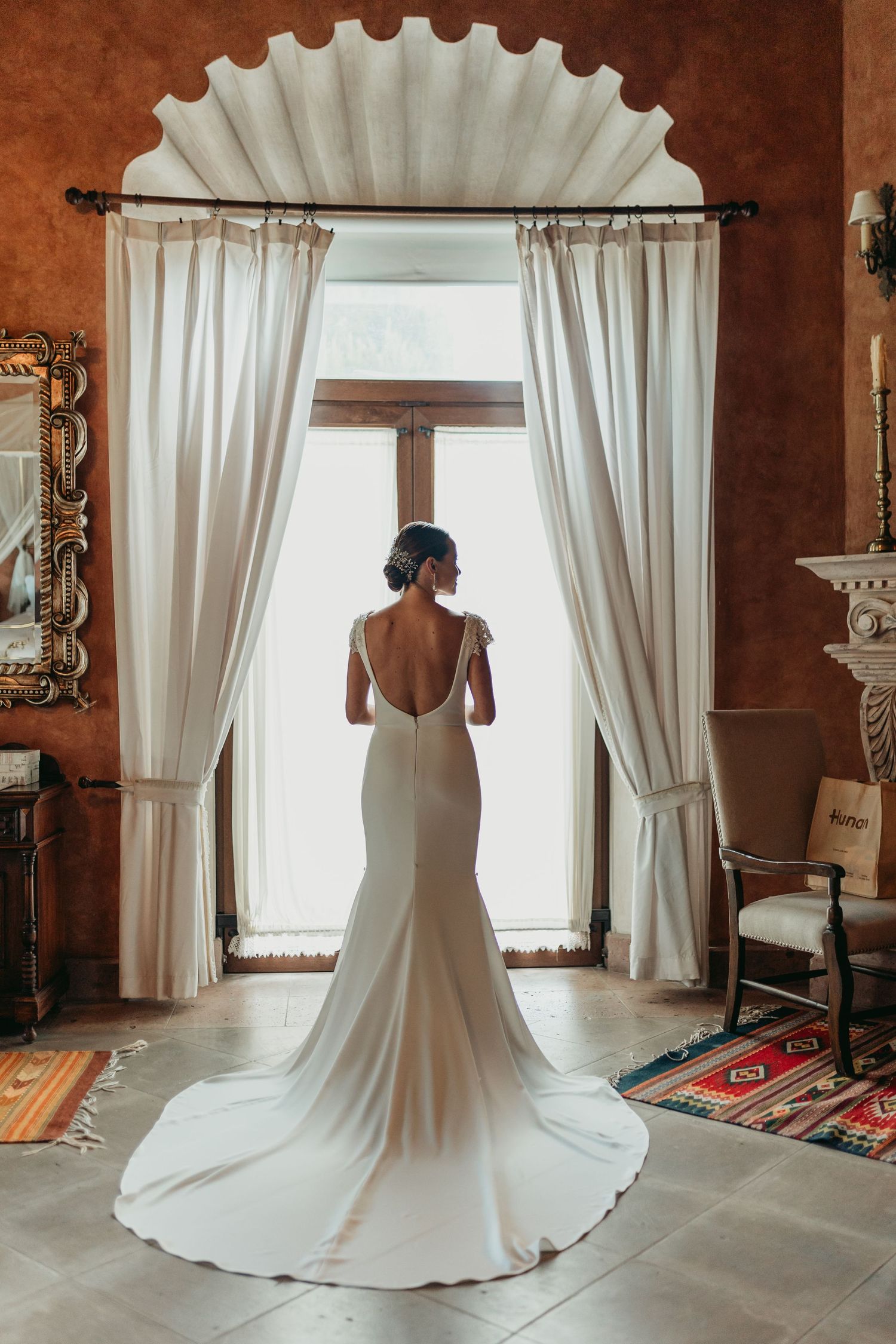 Elegant bridal gown with long train displayed in rustic room with white curtained windows.