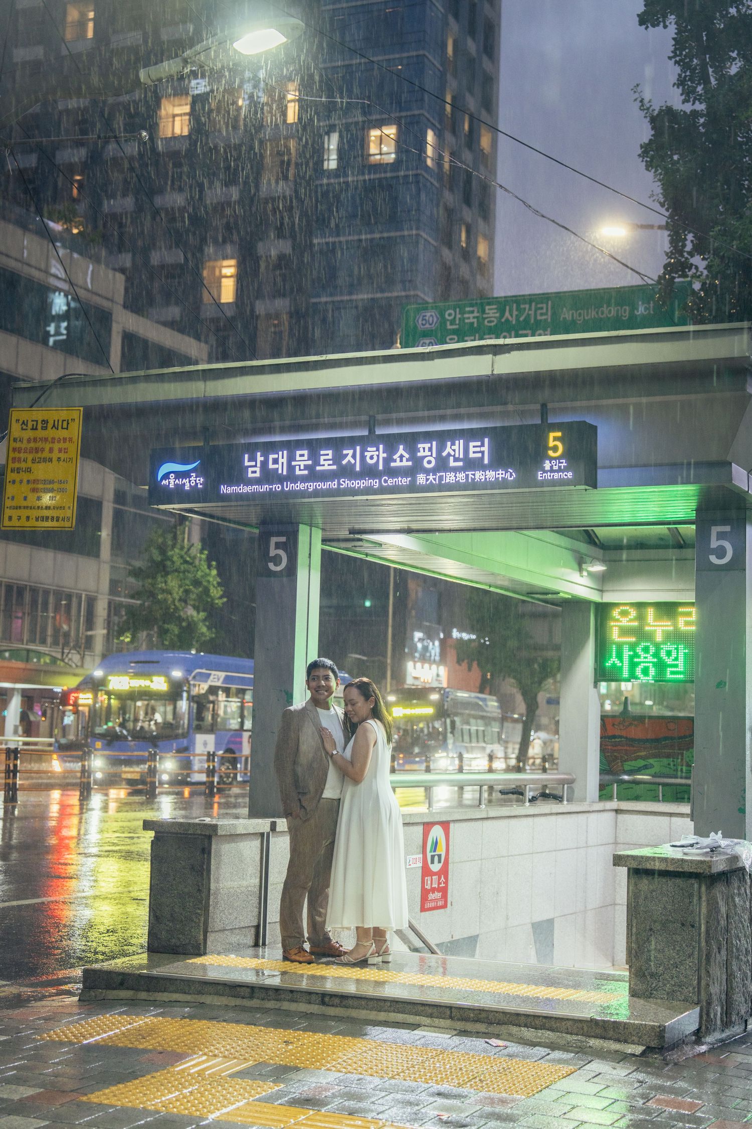 People wait at a brightly lit bus stop shelter in the rain at night in South Korea.