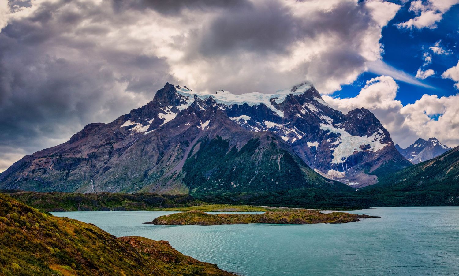 Panoramic view of snow-capped mountain peaks and turquoise lake waters under dramatic cloudy skies in Torres del Paine.