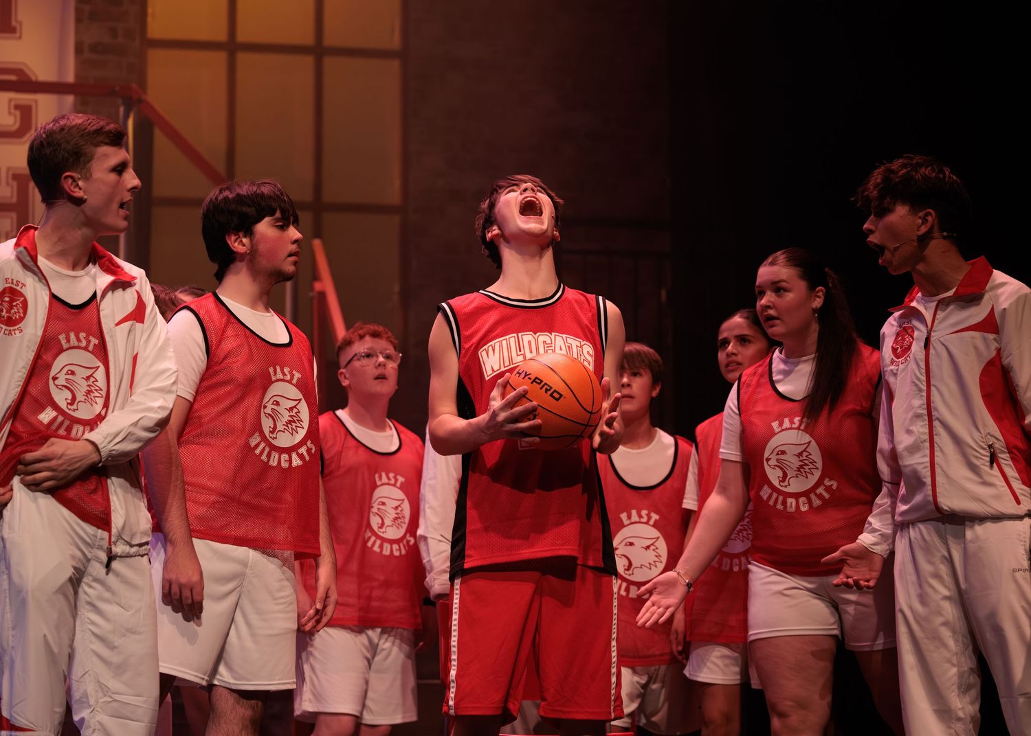 High school performers in varsity jackets and athletic uniforms interact dramatically during a stage production.