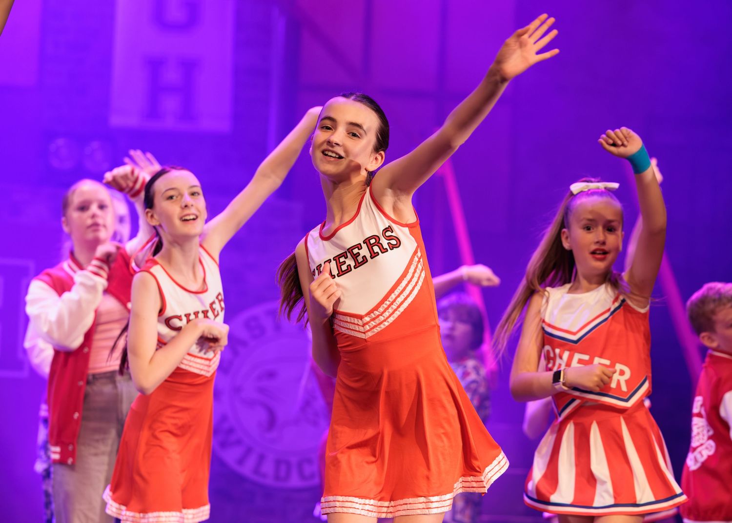 Dancers in red and white cheerleading uniforms perform synchronized movements on stage under purple lighting.