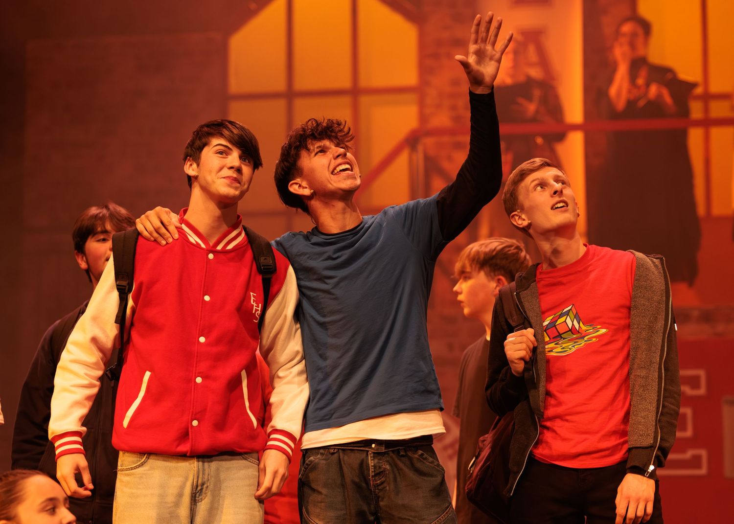 Students in red and white uniforms perform energetically on stage under warm orange lighting during a theatrical show.