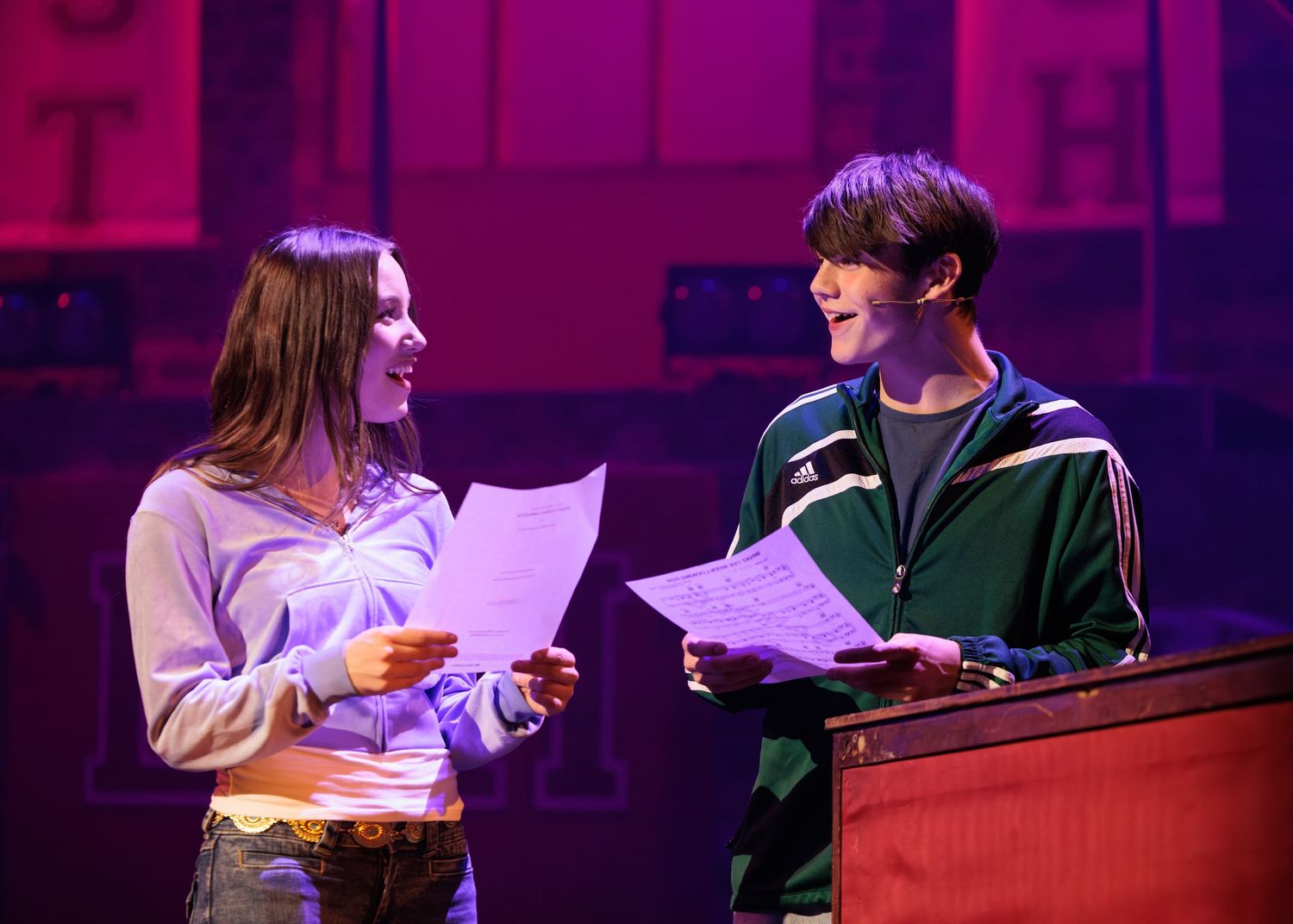 Two performers hold scripts and read together on stage under dramatic purple lighting during a theatrical production.