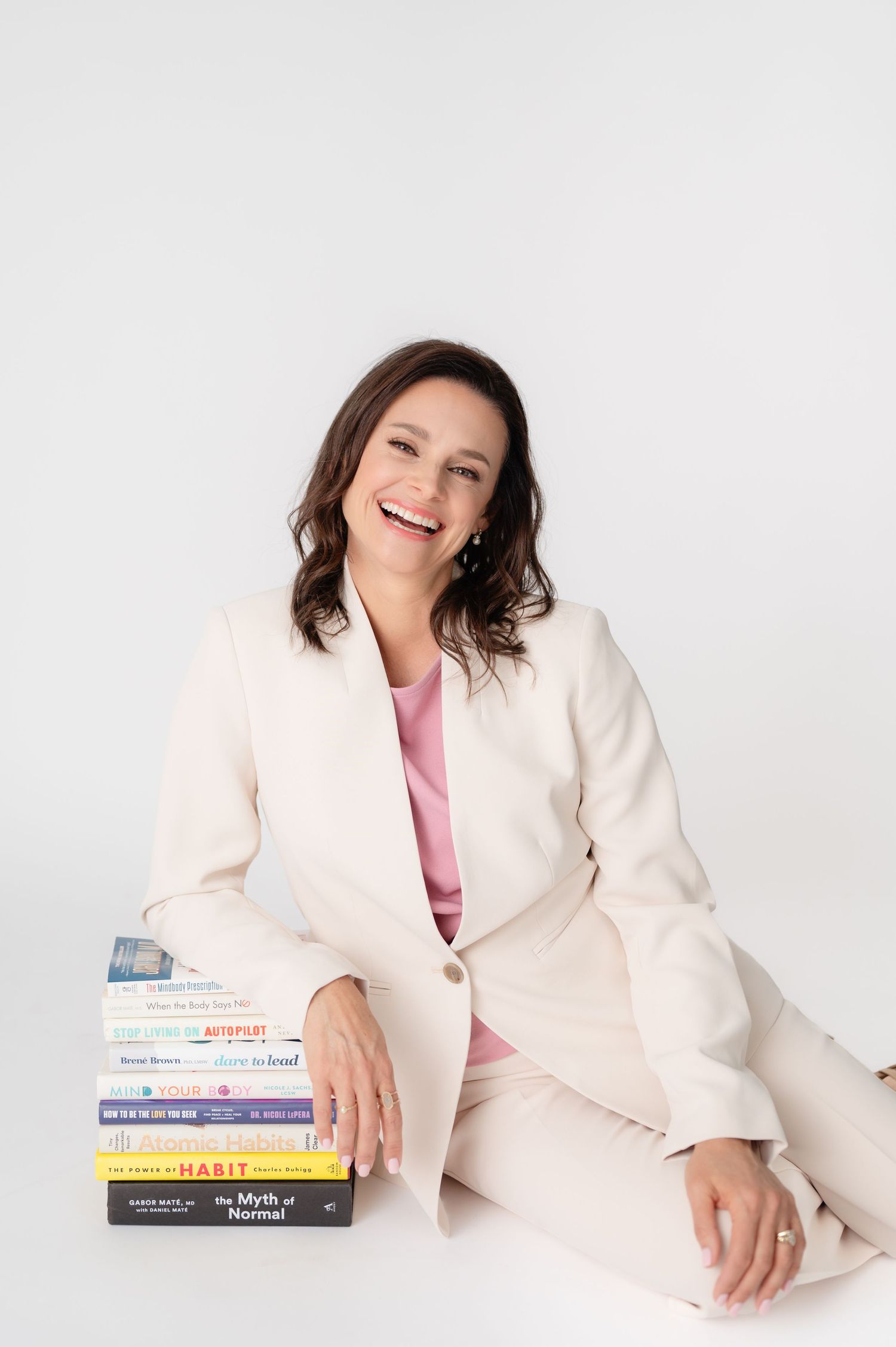 Professional wearing a white blazer and pink top poses with books in a modern studio setting against a light background.