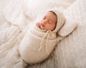 A peaceful sleeping newborn wrapped in cream knit wear on soft white textured blanket.