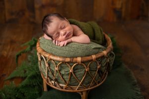 Sleeping newborn nestled in woven bamboo basket with green blanket in rustic setting.