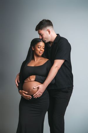 Interracial couple in matching black outfits embracing pregnant belly tenderly in professional studio maternity portrait.