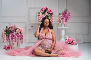 Pregnant woman in pink lace maternity gown sits gracefully surrounded by pink floral arrangements in white studio.