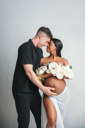 Couple sharing a tender maternity moment, woman in white wrap dress with magnolia flowers, man in black outfit.