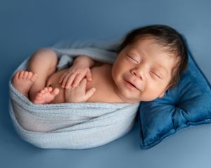 Smiling newborn baby wrapped in light blue swaddle sleeping on blue background with small velvet pillow.