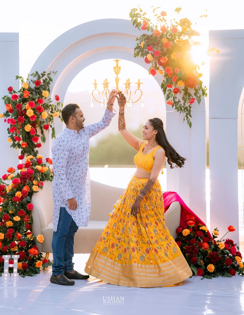 Couple performs romantic dance moves against white archways adorned with vibrant red flowers.