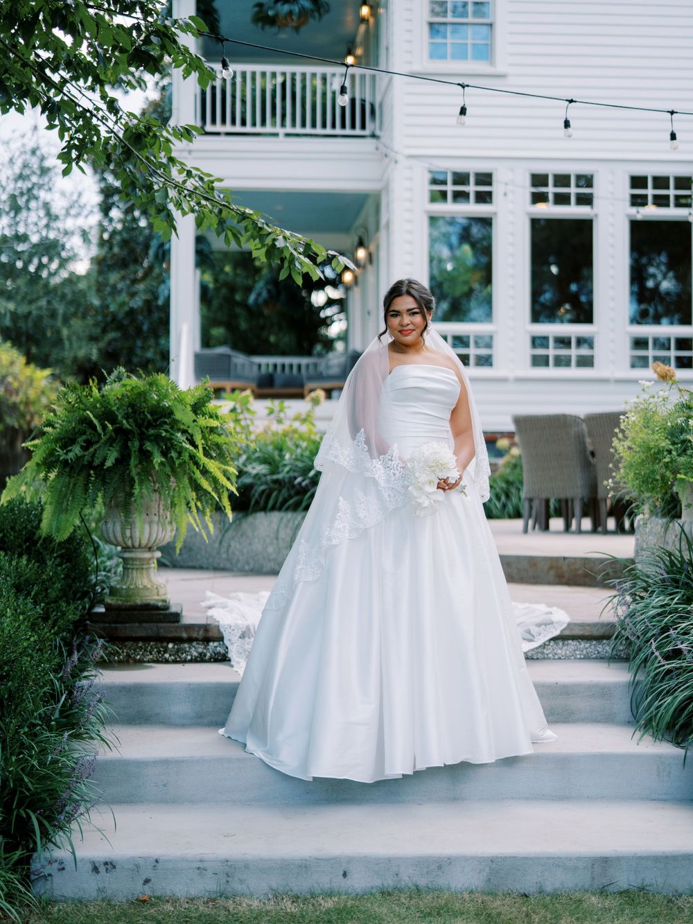 A bride in a flowing white gown stands on a porch surrounded by lush greenery and white columns.
