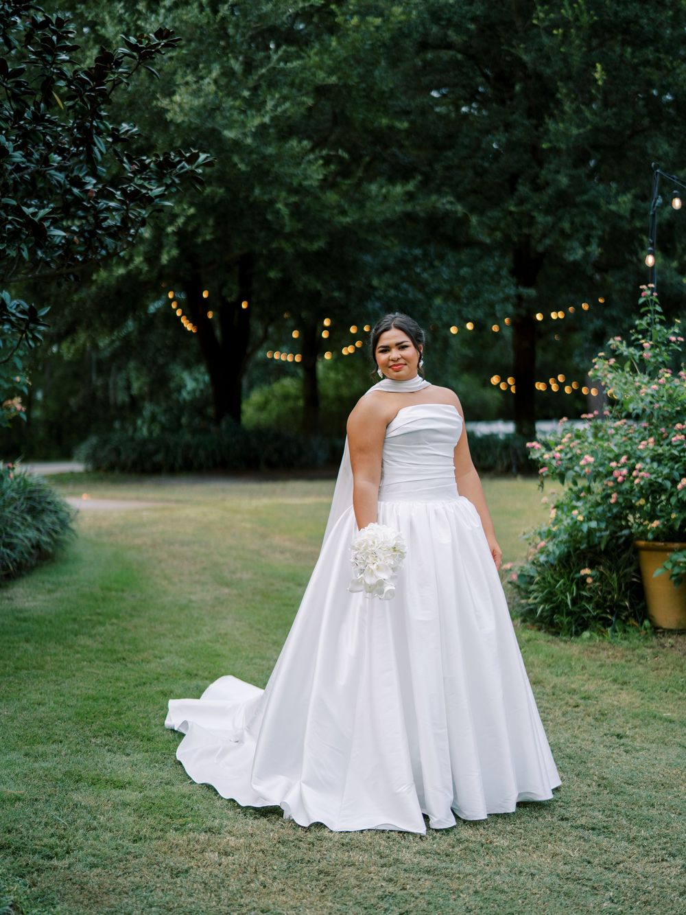 Bride in strapless white wedding gown poses in garden setting with string lights and greenery.