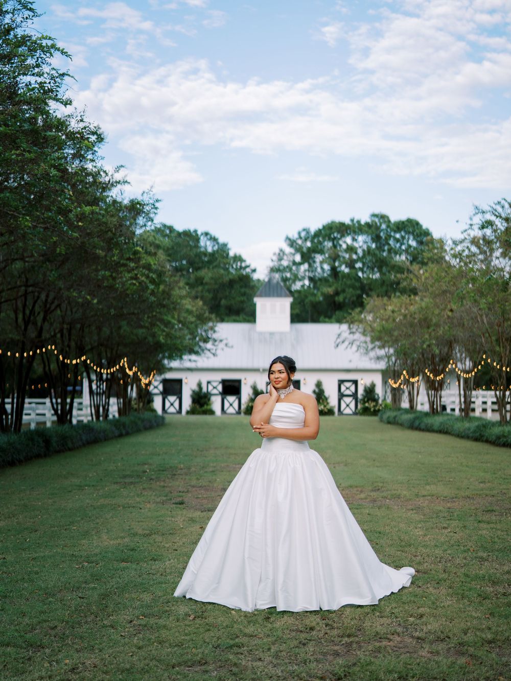 A bride in a flowing white ballgown stands in an outdoor garden venue with string lights and white columns.