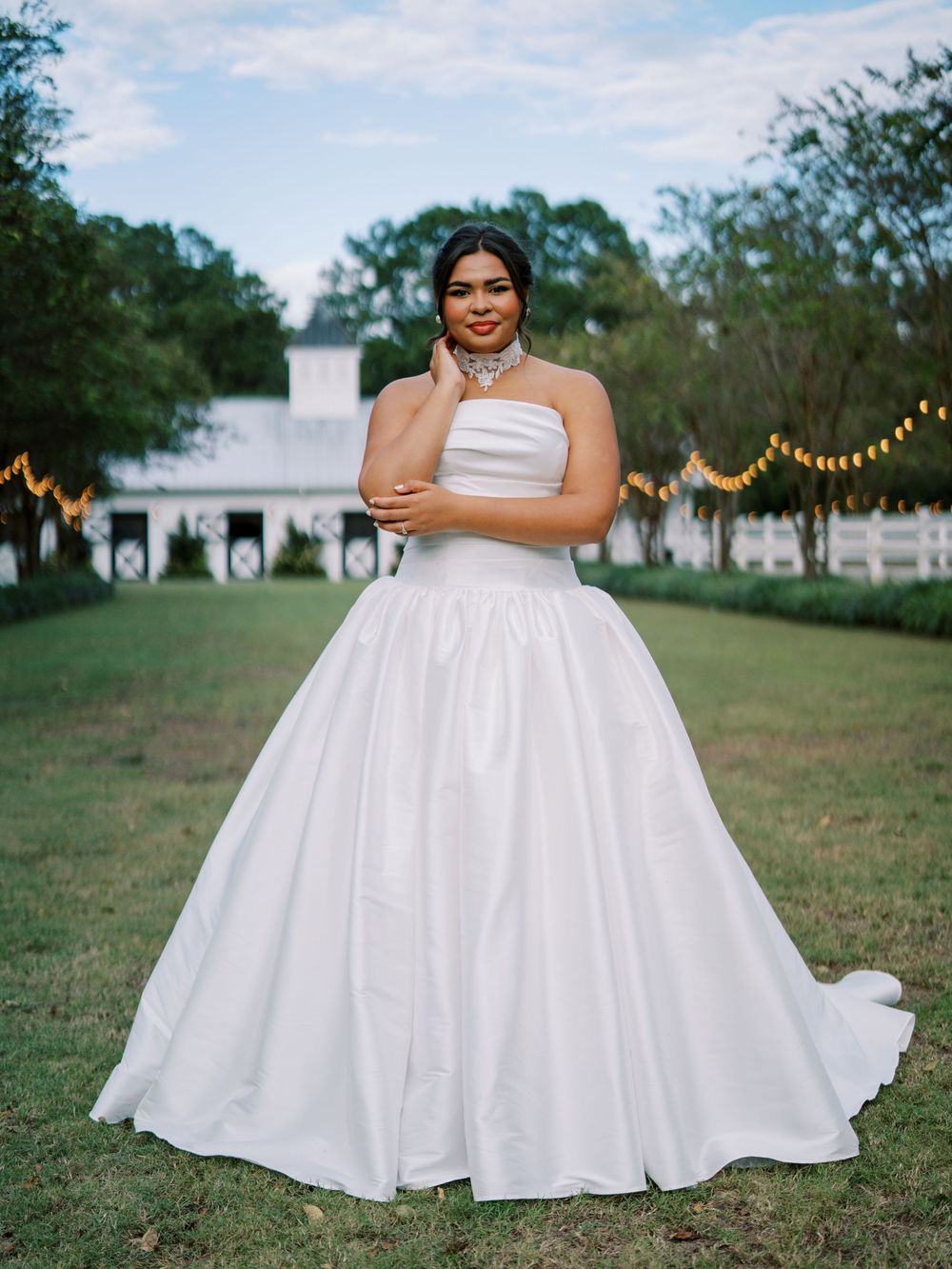 A bride in a flowing white ball gown poses in a scenic outdoor setting with string lights and trees in the background.