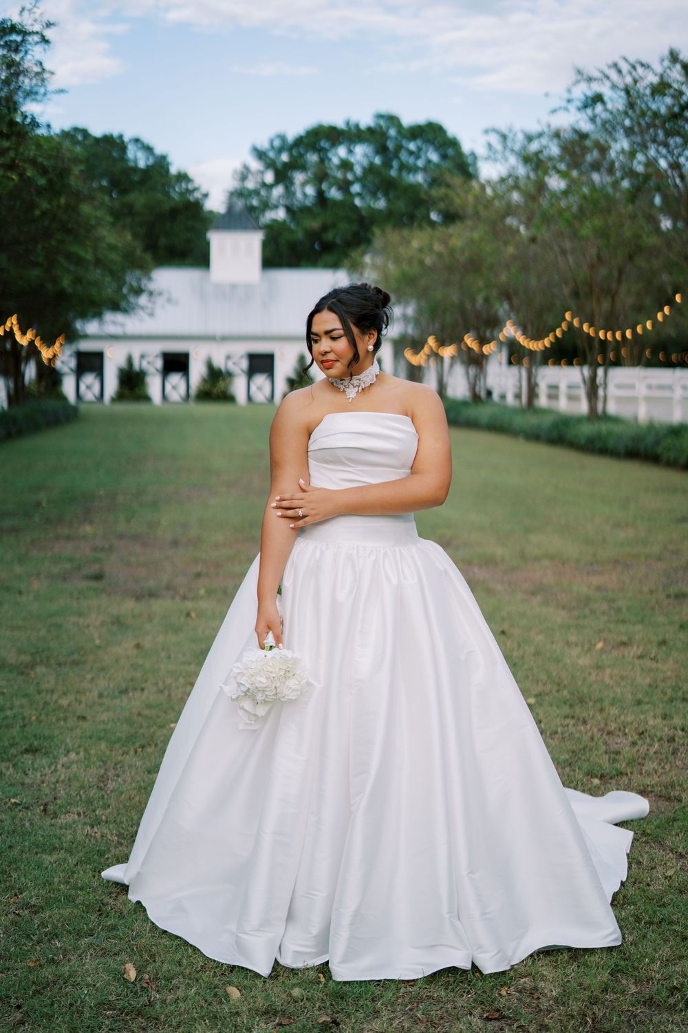 Bride in a classic white ballgown poses gracefully on grassy grounds with string lights in background.