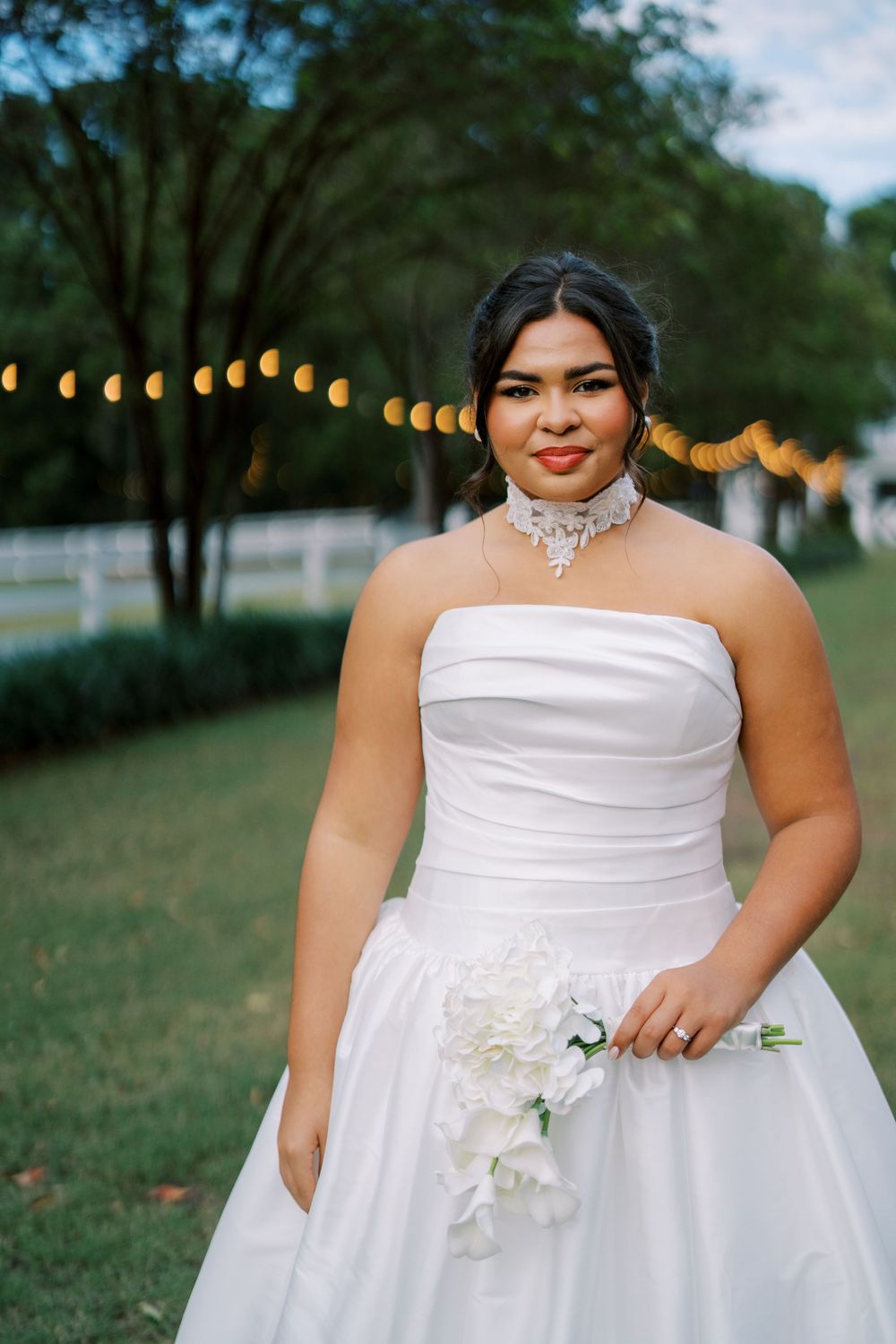 Bride wearing strapless ballgown poses outdoors with string lights and floral details.