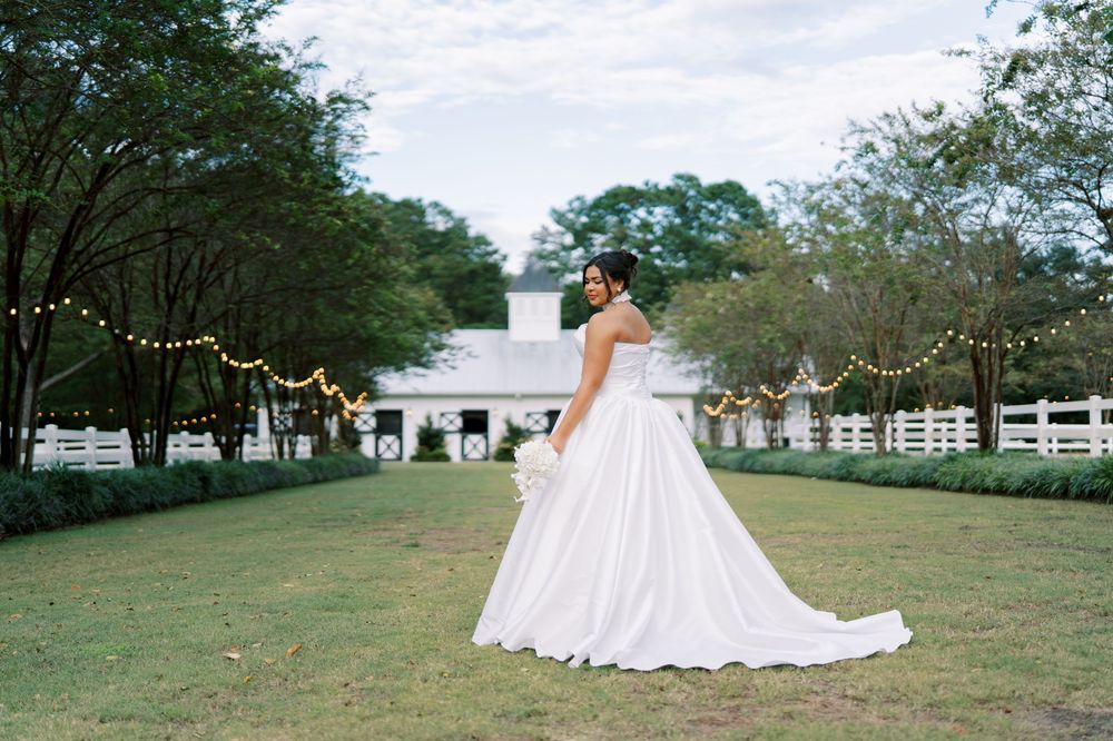 A person in a flowing white wedding dress walks across a scenic green field with white fencing in the background.