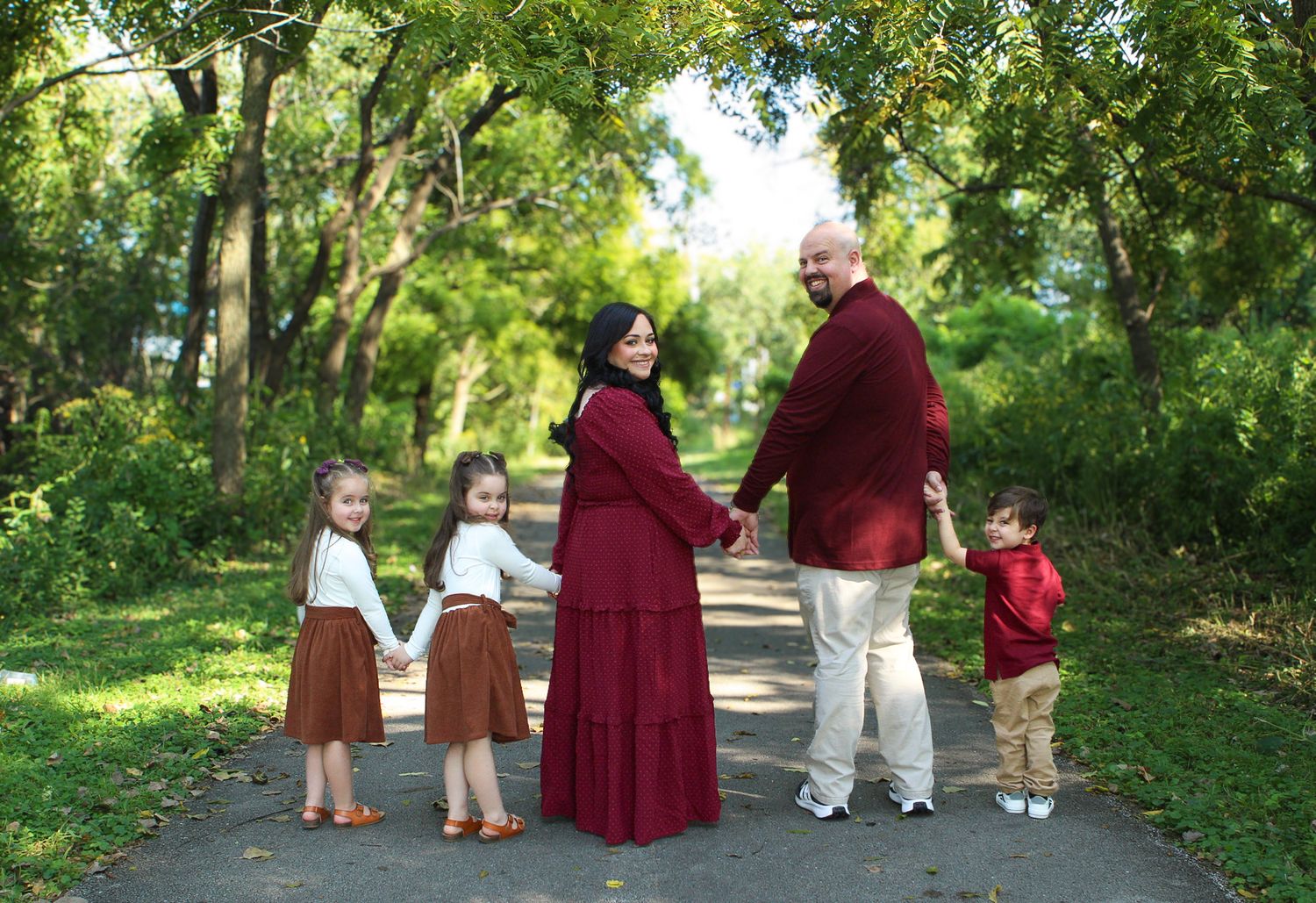 Family portrait session in a lush green park pathway with coordinated burgundy and white outfits.