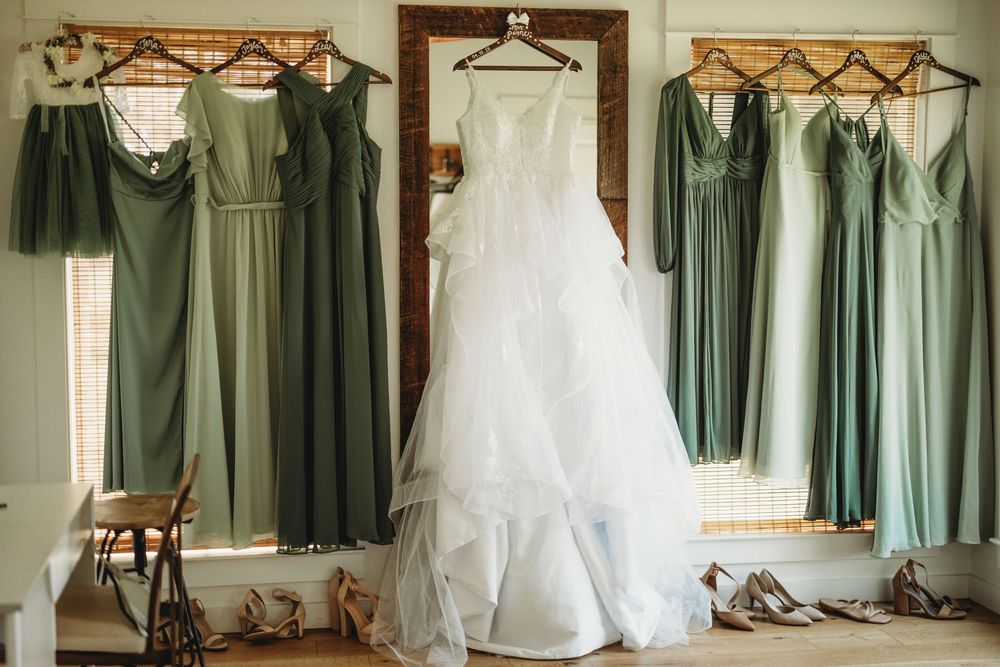 Sage green bridesmaid dresses hang alongside white wedding gown in preparation room.