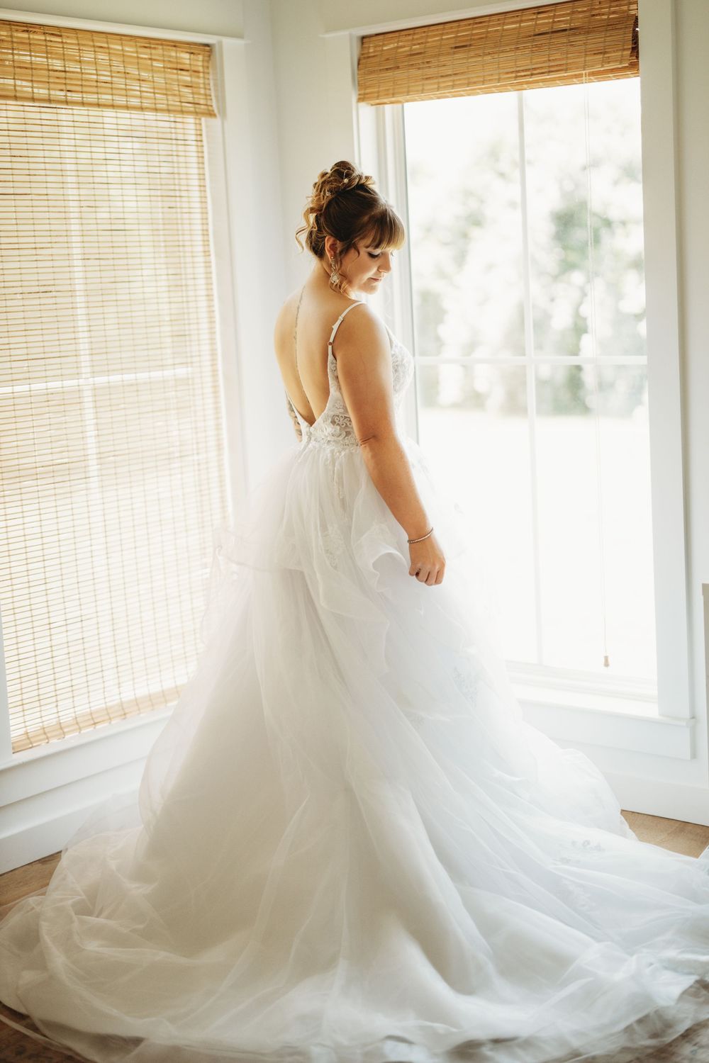 Bride in flowing white wedding gown twirling in sunlit room with bamboo shades.