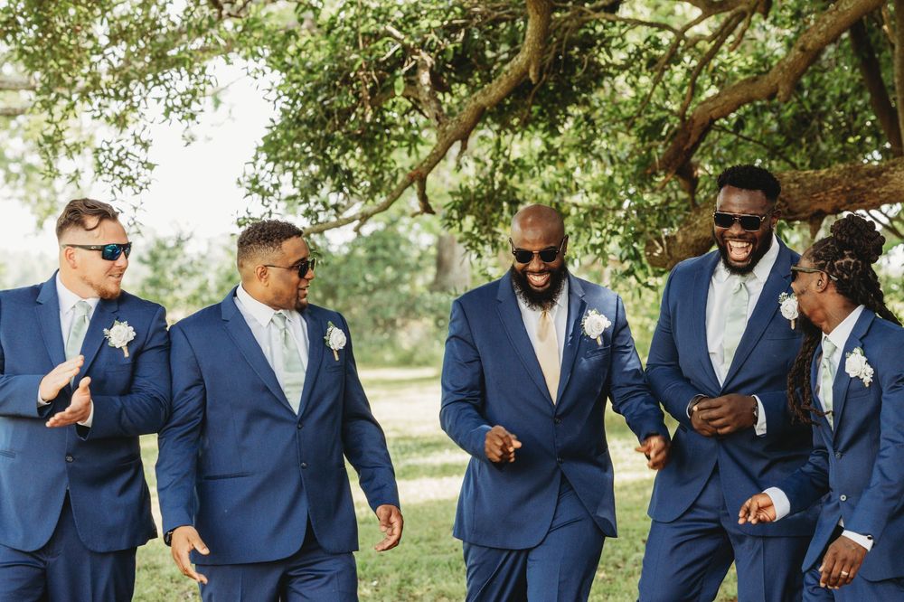 Groomsmen in navy blue suits laugh and smile while standing together outdoors under trees.