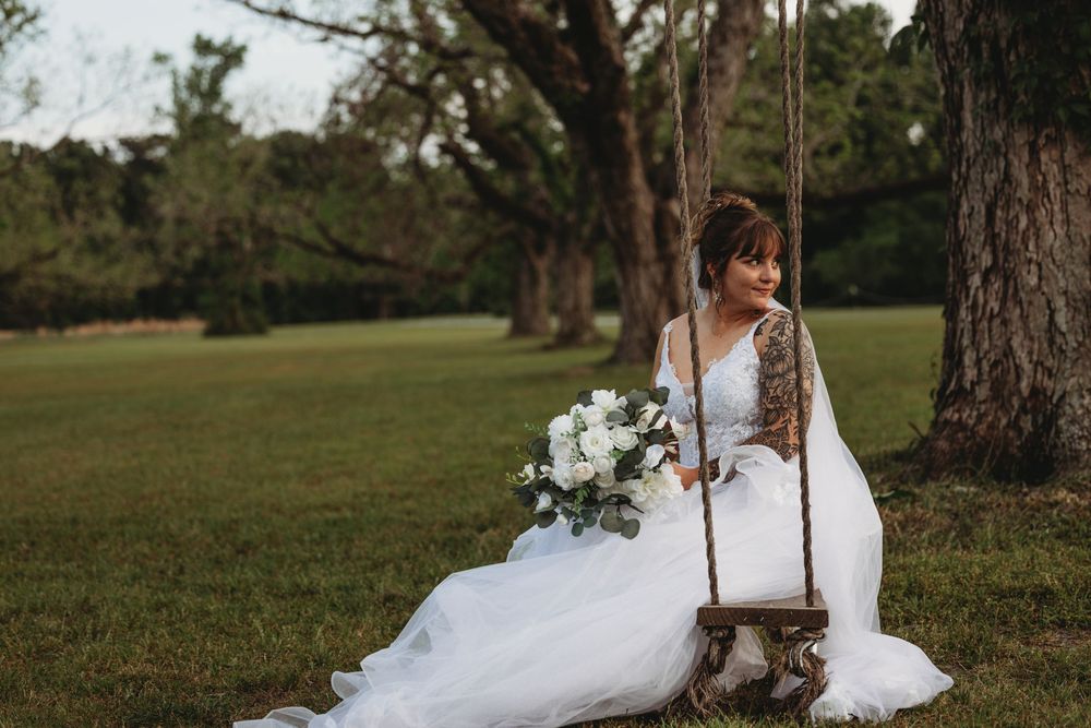 Bride in white dress posing on tree swing in outdoor forest setting.