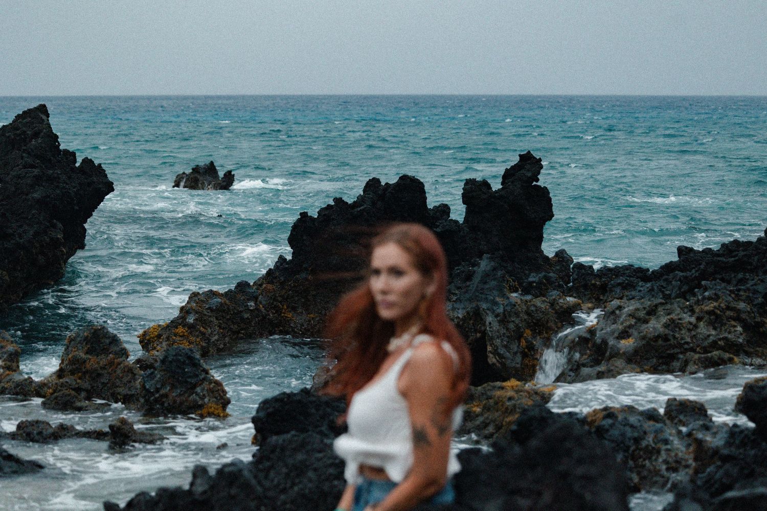 Woman with red hair in white top poses on dark rocky terrain while looking upward at dramatic angle.