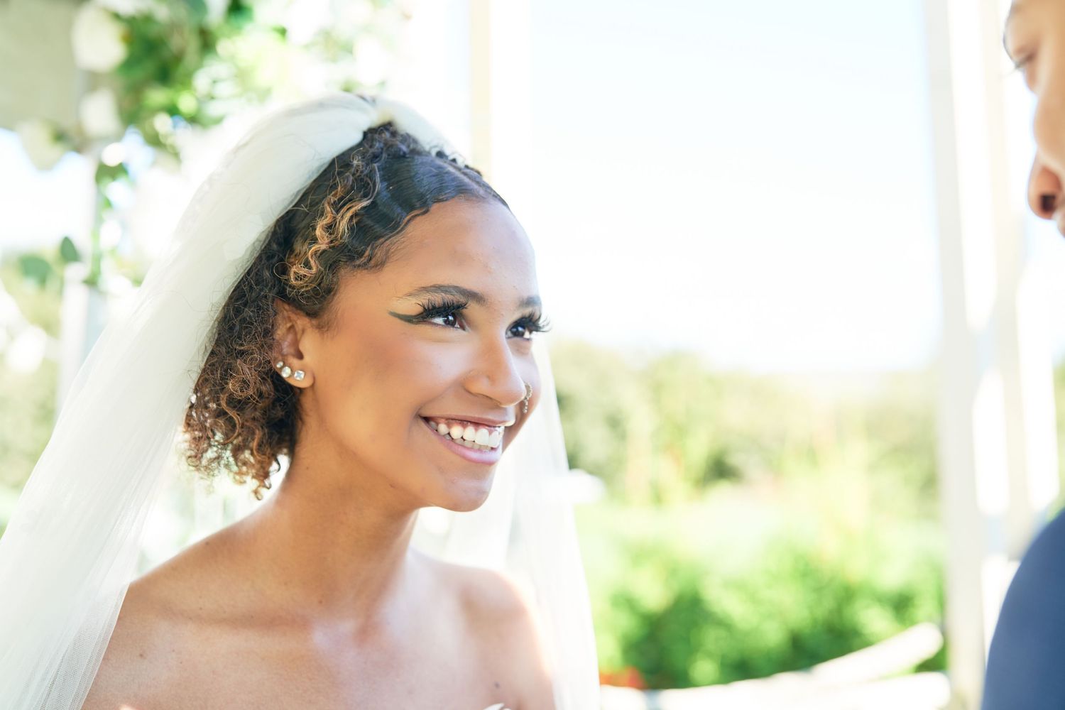A radiant bride wearing a white veil and delicate hair accessories smiles warmly during an outdoor wedding ceremony.