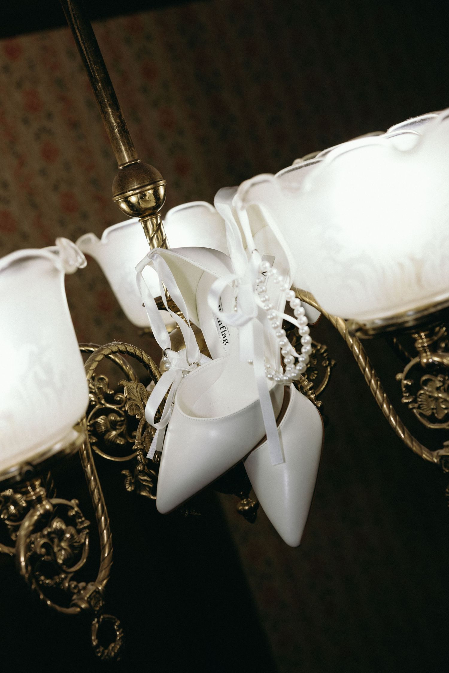 White high heel wedding shoes hanging decoratively from an ornate brass chandelier with frosted glass shades East Coast wedding photographer