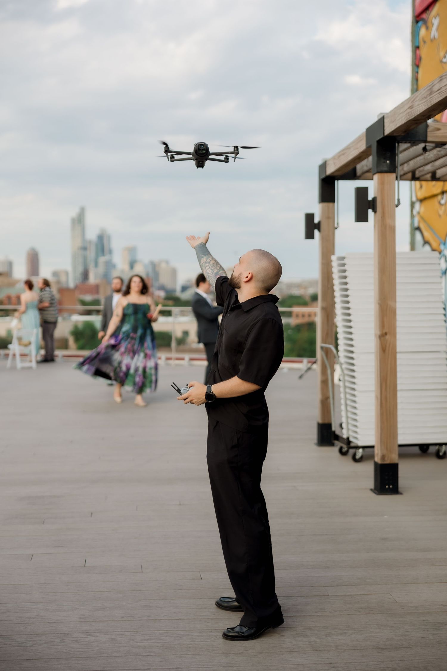 A person in black clothing operates a drone on a rooftop deck with the city skyline visible in the background.