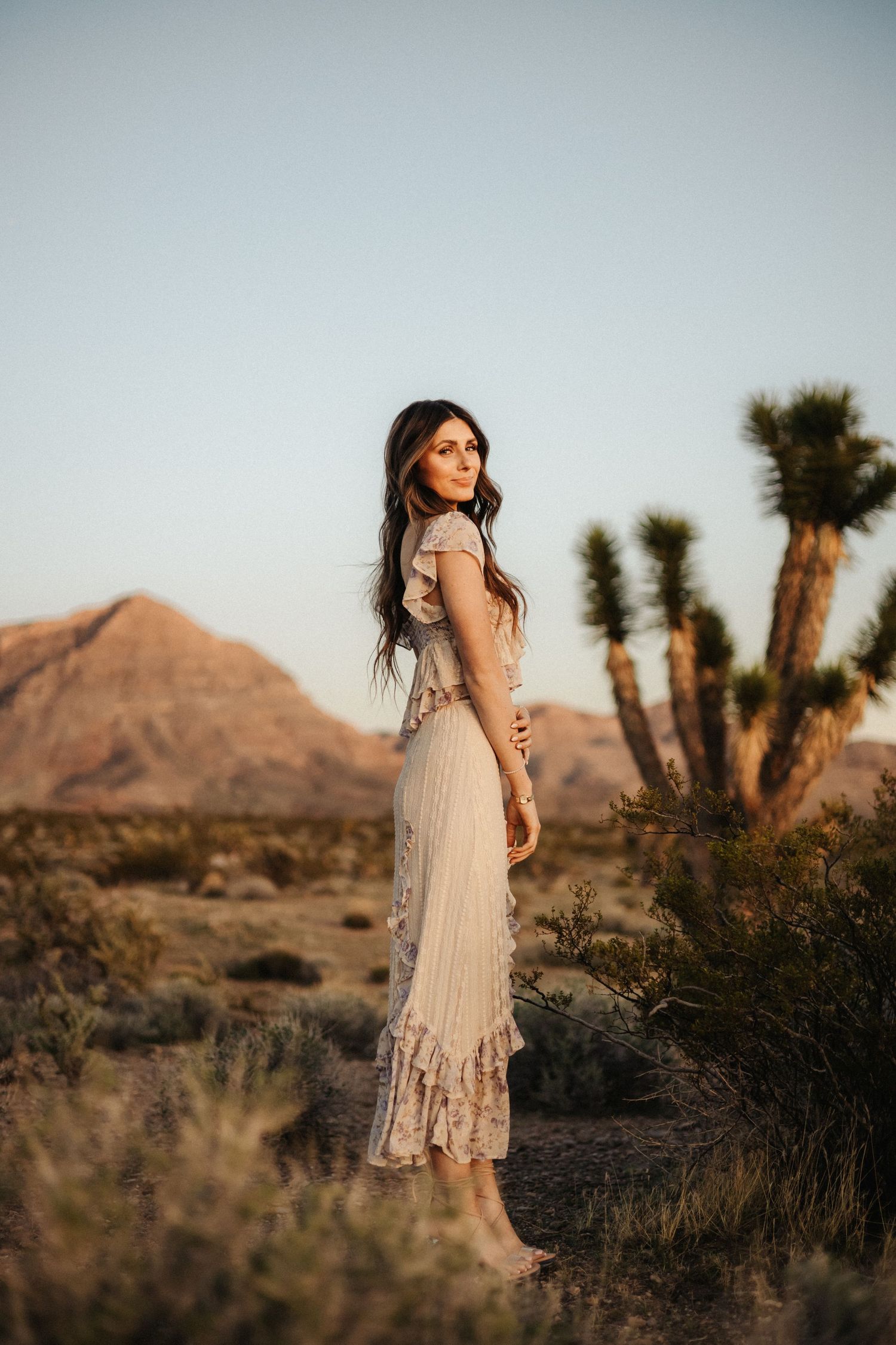 A woman in a flowing white dress poses against a desert landscape with Joshua trees and mountains at sunset.