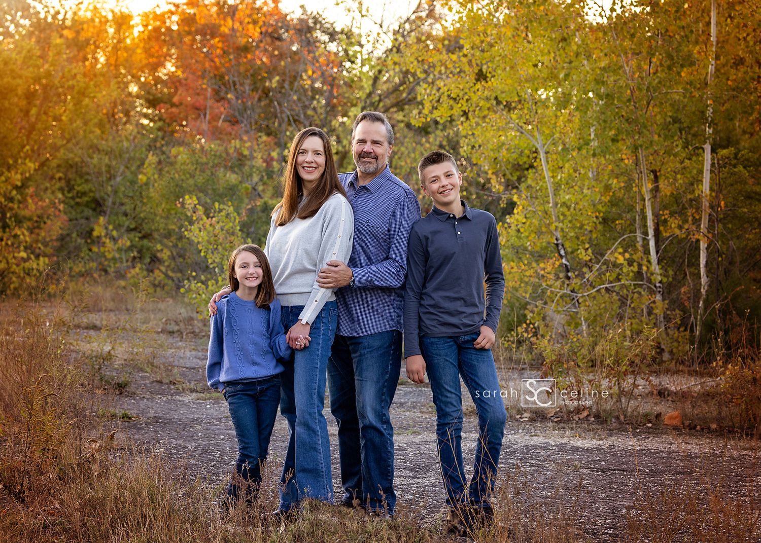 A group of four people standing together on a dirt path surrounded by autumn trees at sunset.