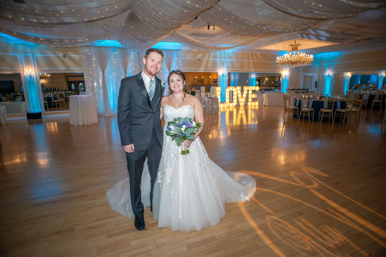Wedding couple poses in a blue-lit ballroom with elegant decor and polished dance floor.