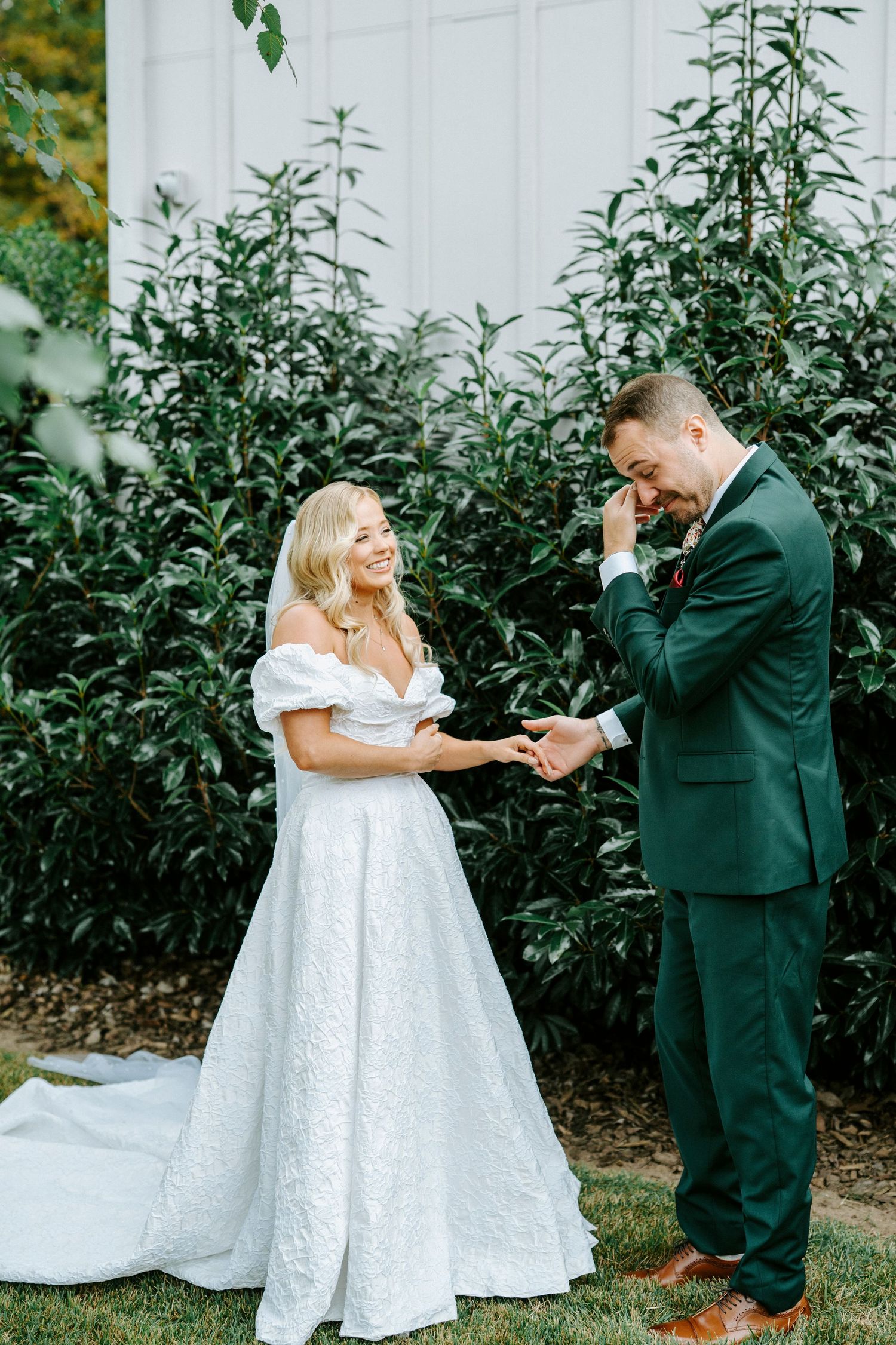 A bride and groom share a playful moment during their first look in a garden setting with lush greenery.