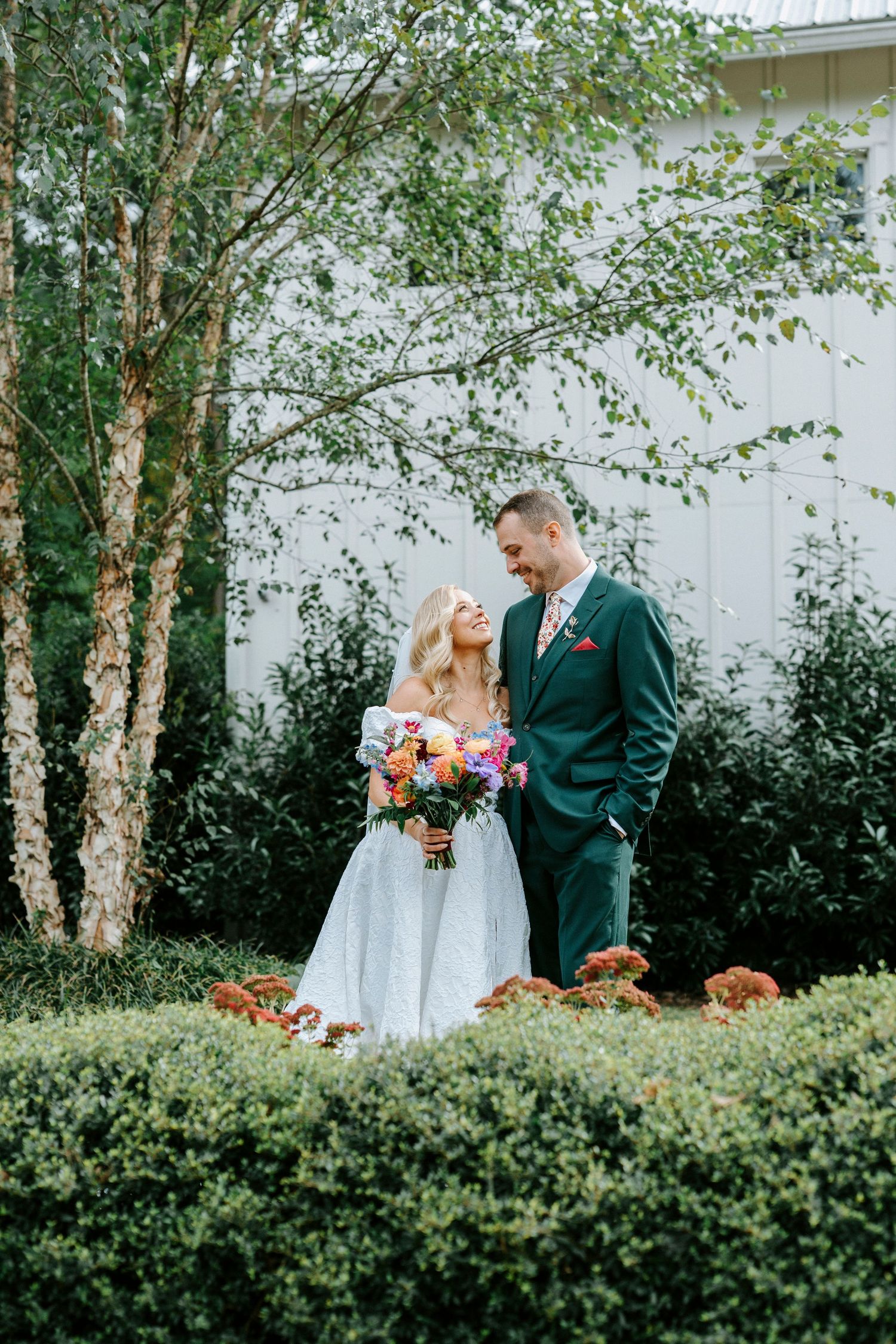 A couple shares a romantic moment in their wedding attire against a white barn backdrop with autumn pumpkins and birch trees.