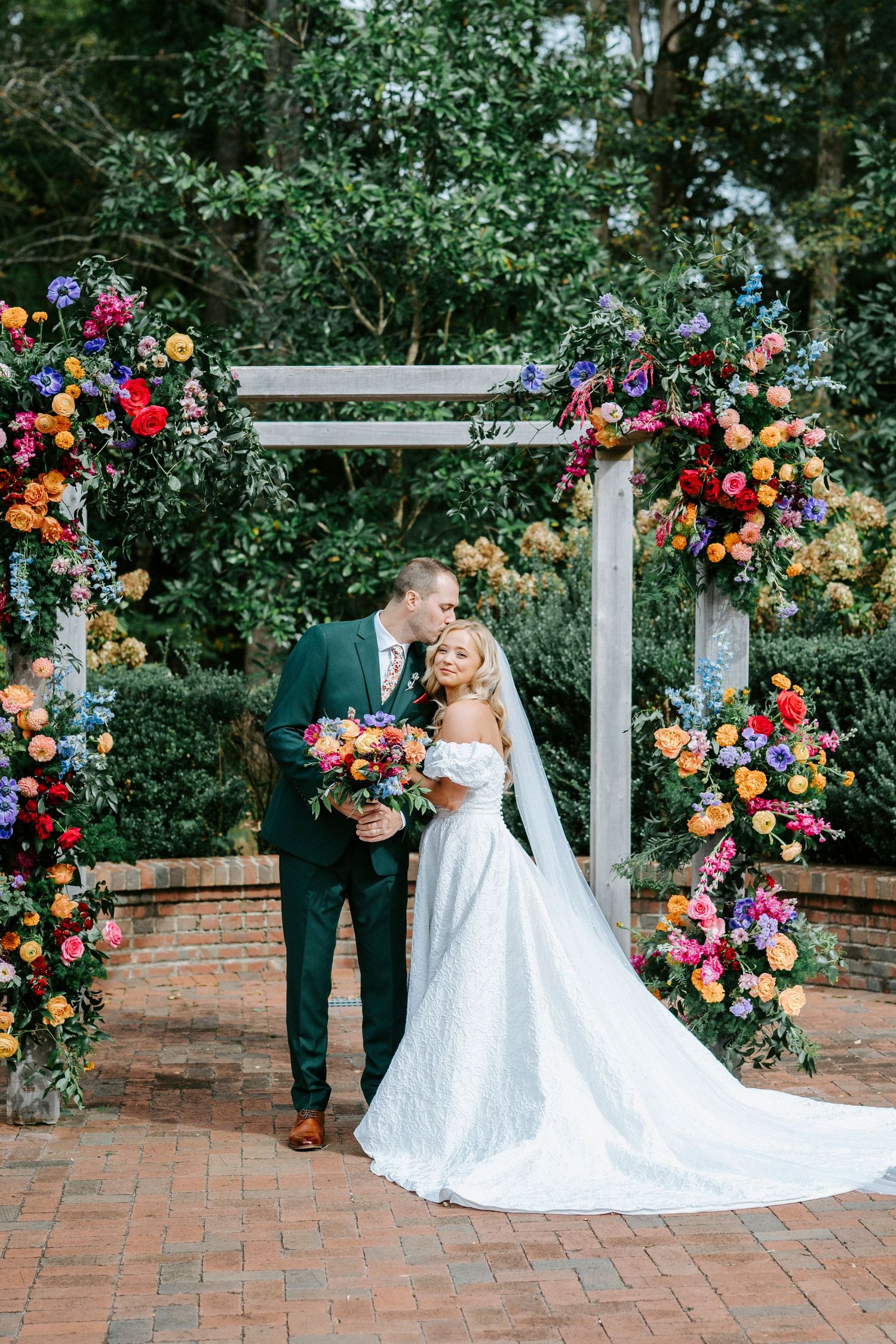 Wedding couple poses under floral arch with vibrant roses and greenery on brick patio during outdoor ceremony.