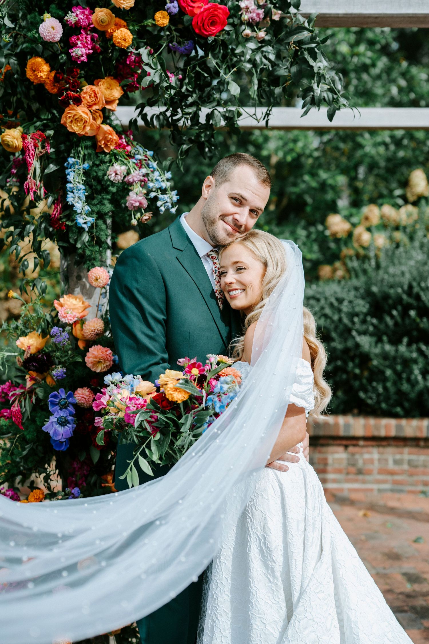 A romantic wedding photo under a colorful floral arch with orange, pink and blue blooms in a garden setting.